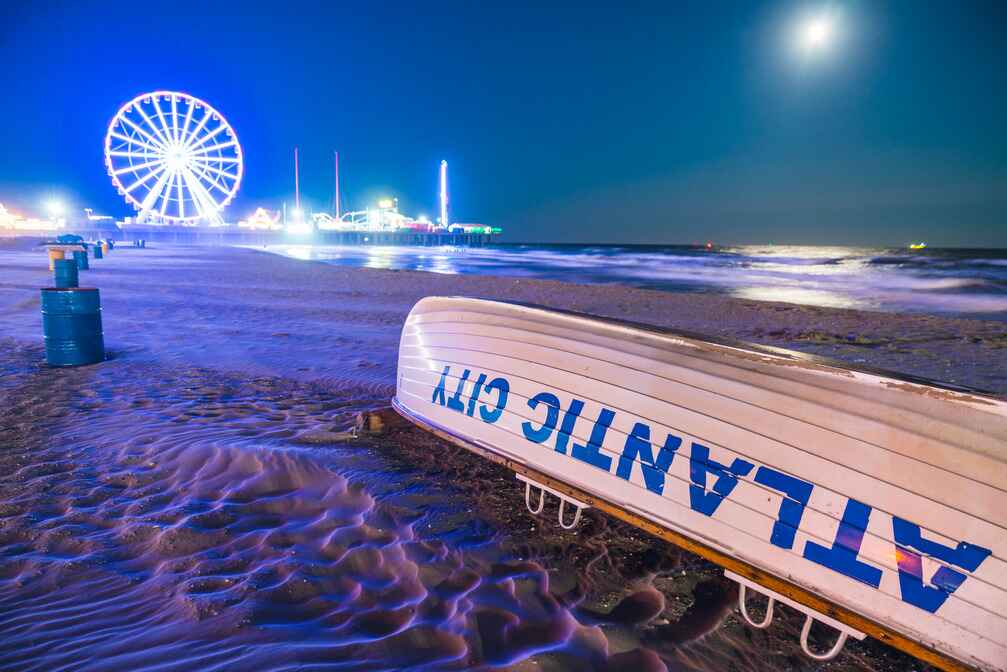 A vibrant nighttime beach scene showcases a Ferris wheel glowing in neon lights, with an overturned lifeguard boat labeled "Atlantic City" resting on the sand in South Jersey, New Jersey.