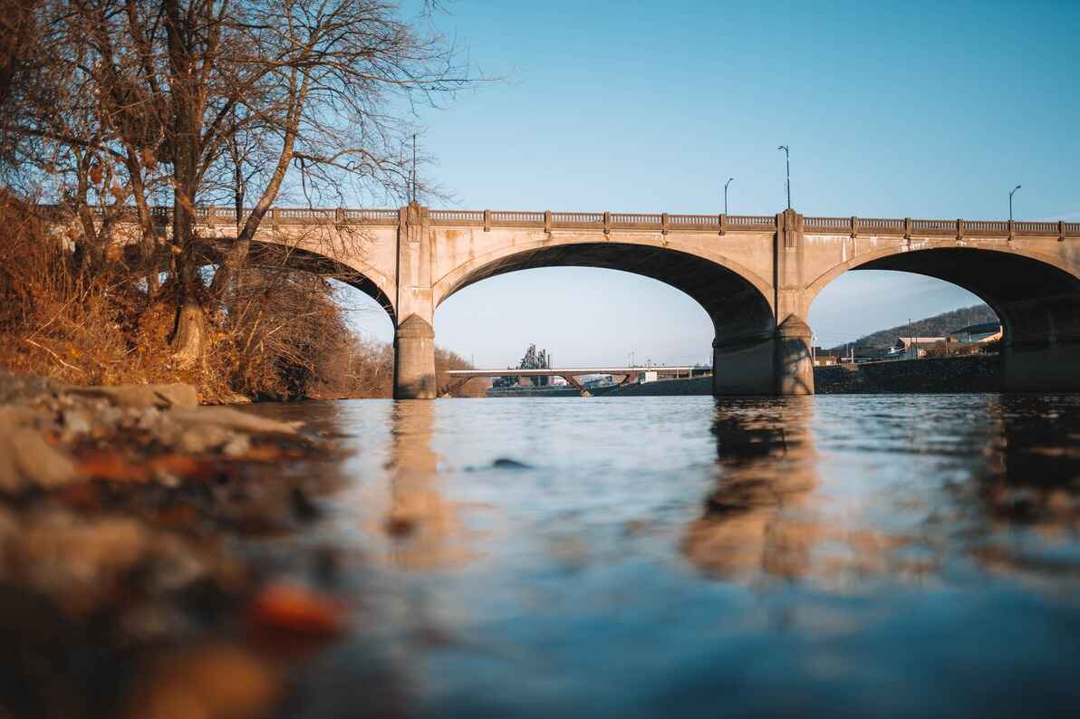 A concrete arch bridge spans a calm river in Lehigh County, with its reflection in the water, framed by leafless trees under a clear blue sky.