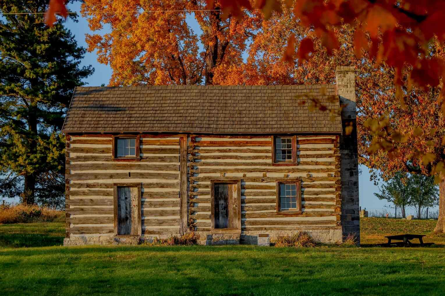 A historic log house called the Flohr House with a stone chimney stands in a grassy field, framed by vibrant autumn leaves in Wythe County, Virginia.