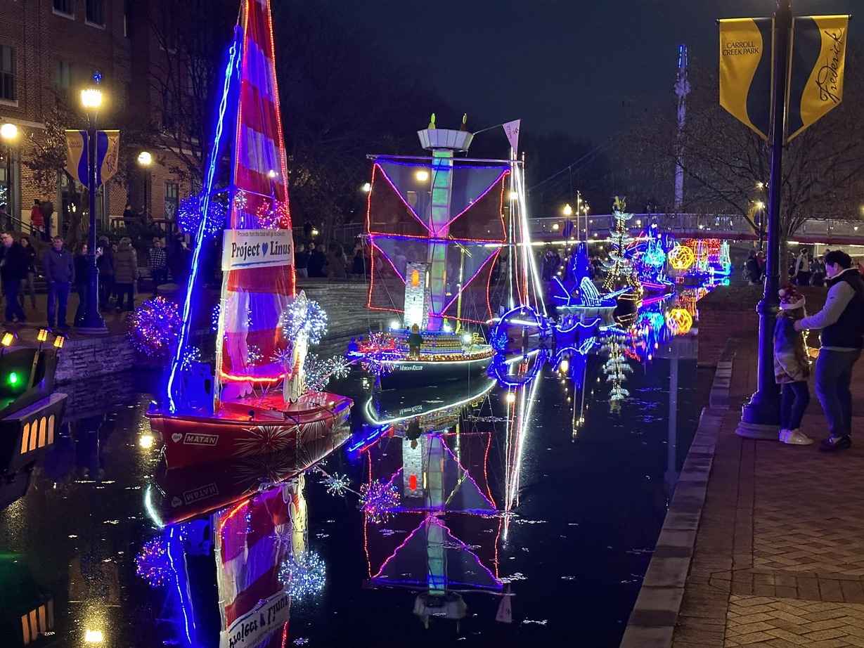 Illuminated boats decorated with colorful lights reflecting on the water during a festive night parade at Carroll Creek Park, Frederick County, Maryland.