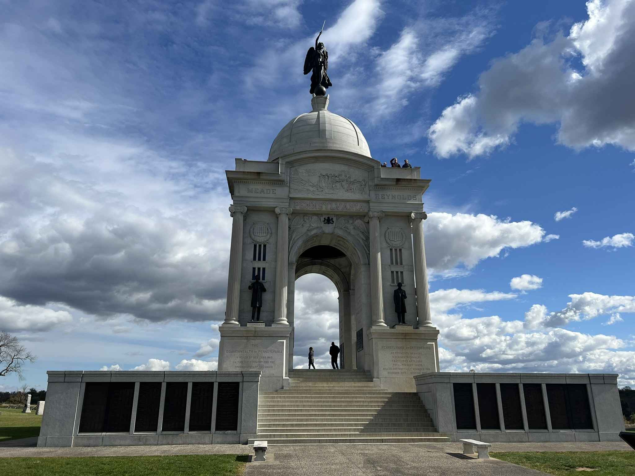 The Pennsylvania Memorial at Gettysburg, a large white marble structure with an angel statue on top, framed by a bright blue sky and scattered clouds.