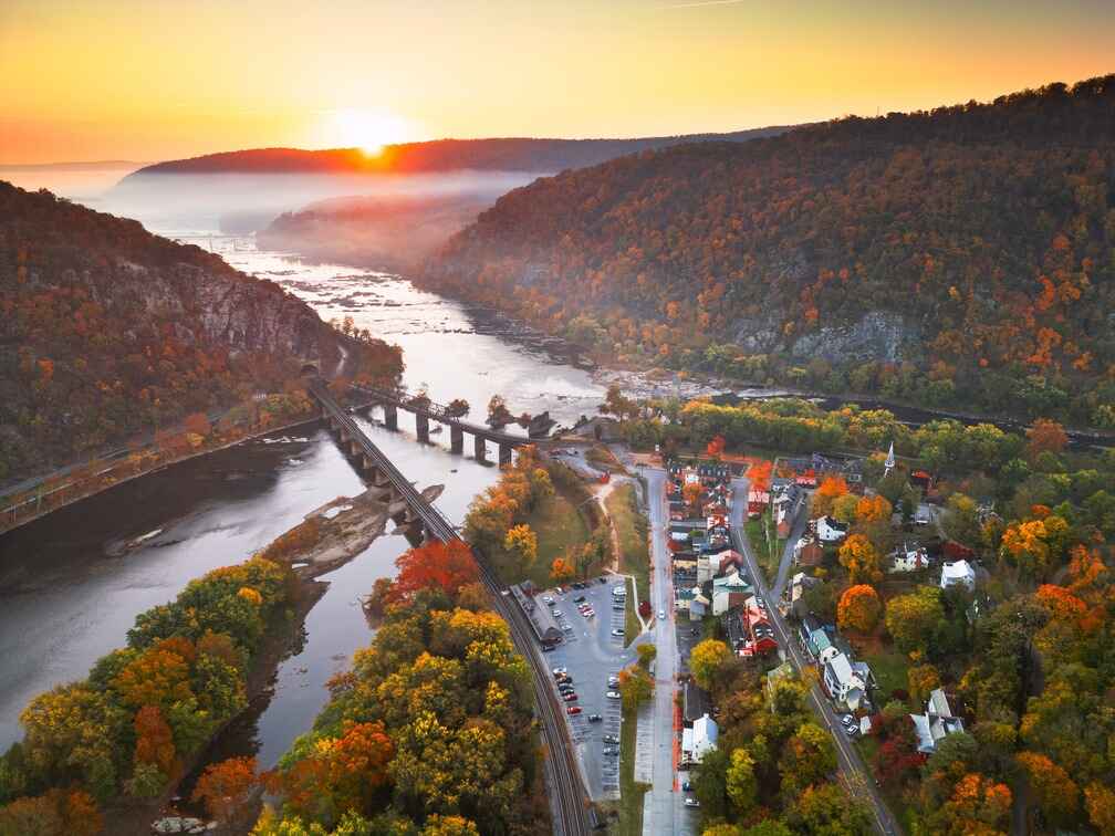 An aerial view of Harpers Ferry at sunrise, showing a small historic town nestled between two rivers and surrounded by misty, autumn-colored mountains in Jefferson County, West Virginia.