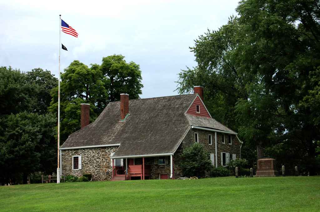 A historic stone house called the Hasbrouck House with a red roof and an American flag stands on a green lawn, surrounded by large trees in Orange County, New York.