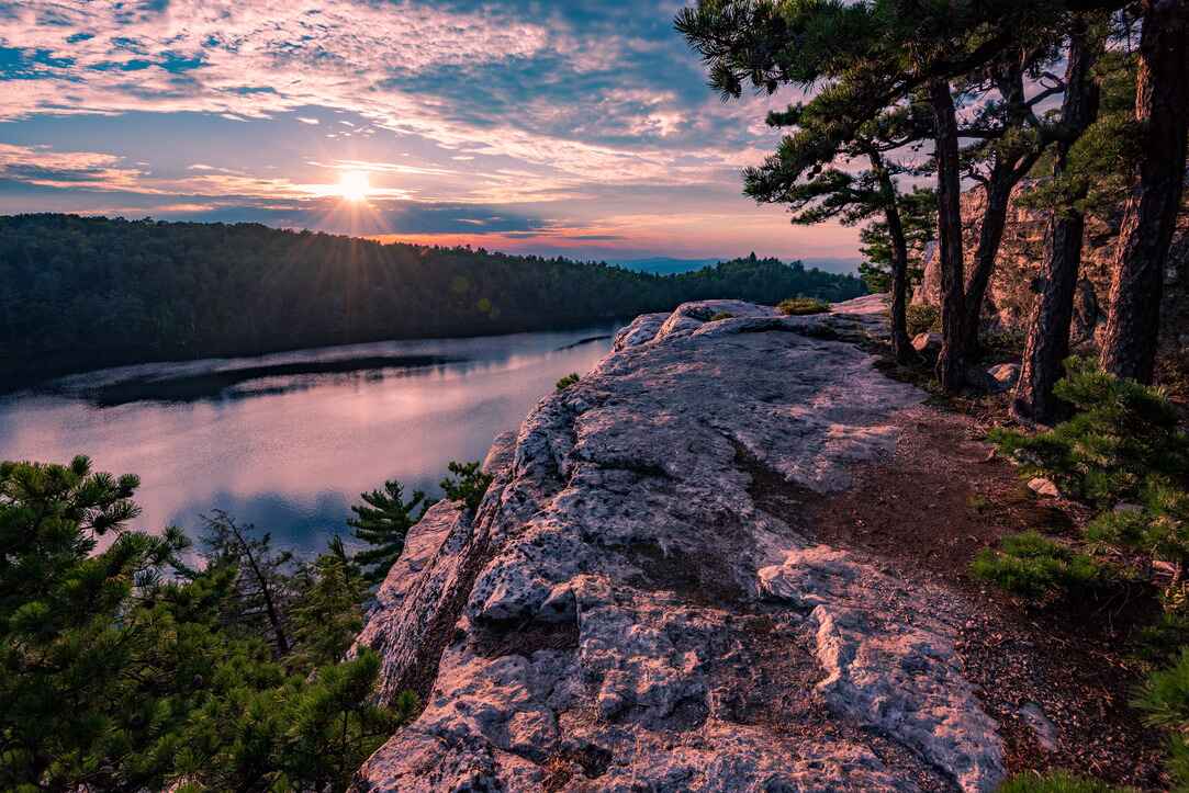 A rocky cliffside overlooks Lake Minnewaska at sunset, with pine trees and a colorful sky reflecting on the water in Minnewaska State Park.