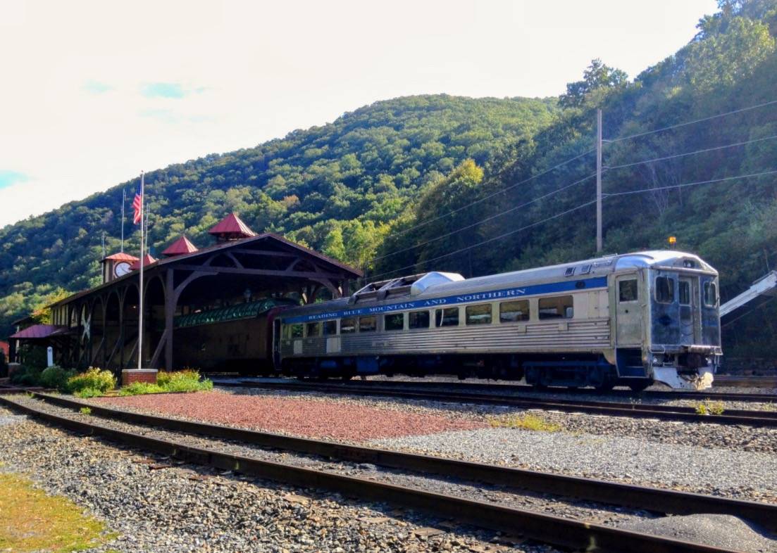 A Reading Blue Mountain and Northern Railroad train car sits on the tracks near a wooden station with red-roofed towers, surrounded by green mountains in Schuylkill County, Pennsylvania.