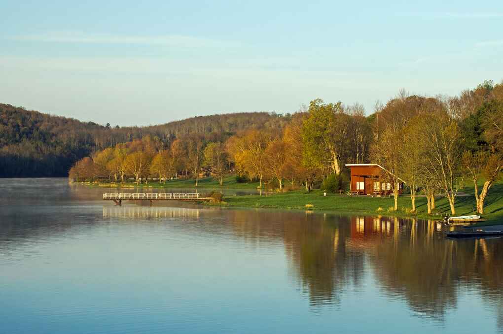 A calm lake reflects the early morning light in Lackawanna County, Pennsylvania, with trees and rolling hills lining the shore.