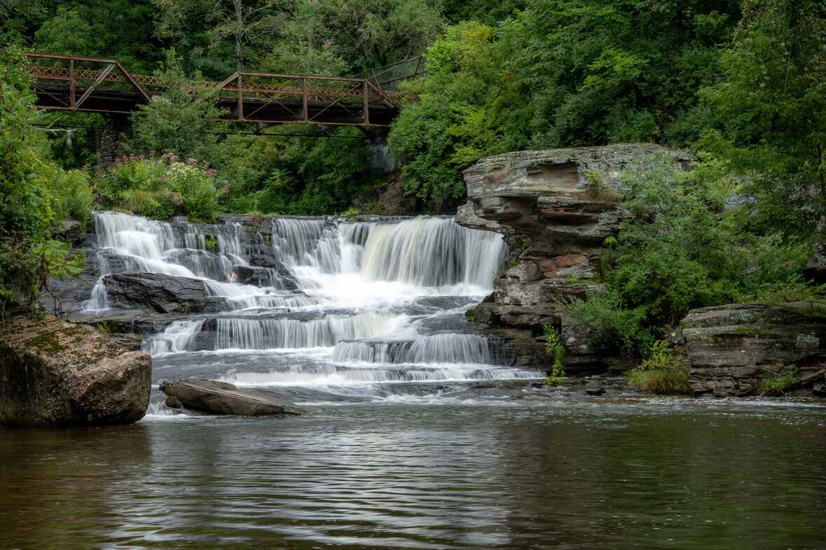 A cascading waterfall surrounded by lush greenery, with a rustic metal bridge spanning the top of the falls in Honesdale in Wayne County, PA.