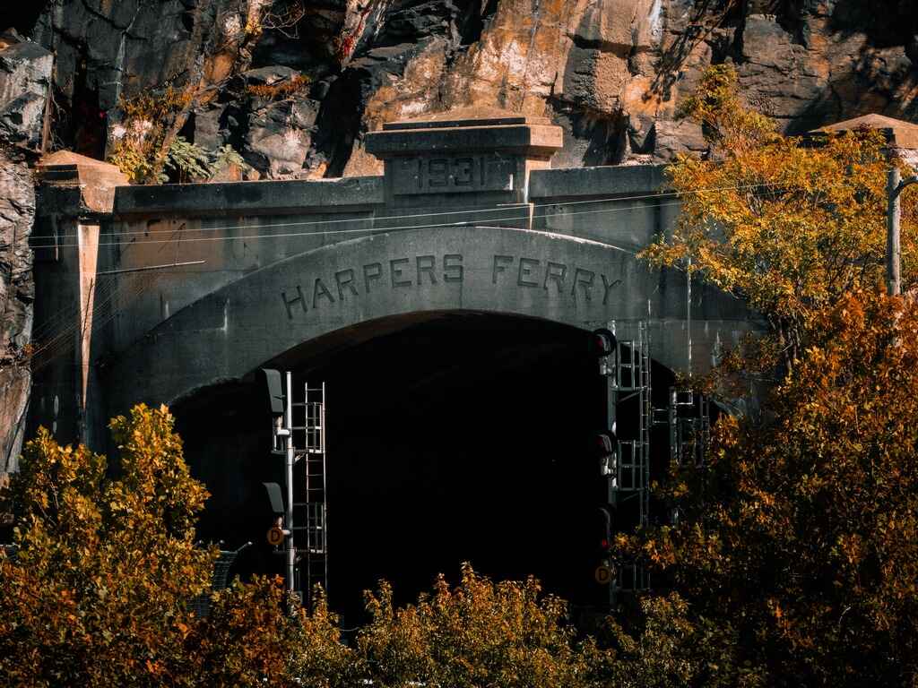 A stone tunnel carved into a mountainside, with the words "Harpers Ferry" engraved above the dark railway entrance, framed by golden autumn foliage in Jefferson County, West Virginia.