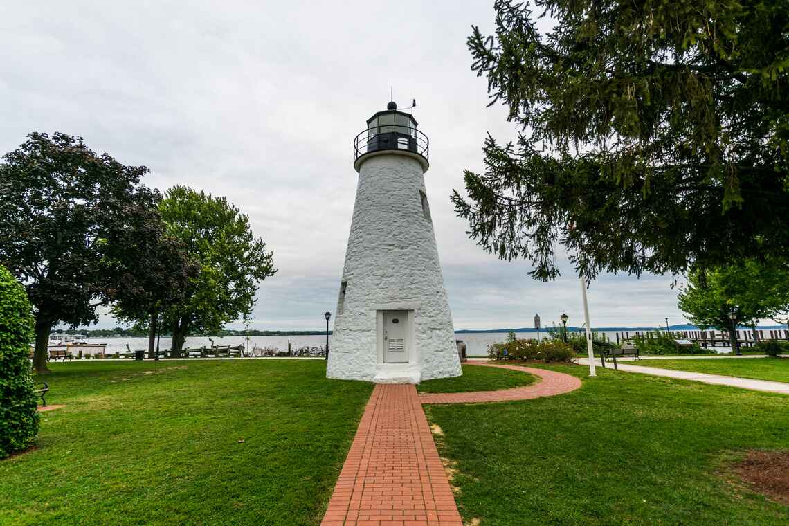A historic white lighthouse standing at the end of a red brick pathway, surrounded by lush green grass and trees in Havre de Grace, Harford County, Maryland.