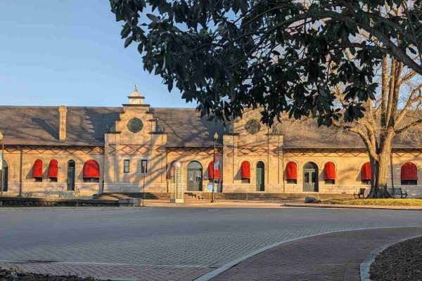 Historic train station in Danville, Virginia, with beige stone walls, arched windows with red awnings, and a paved plaza with trees.