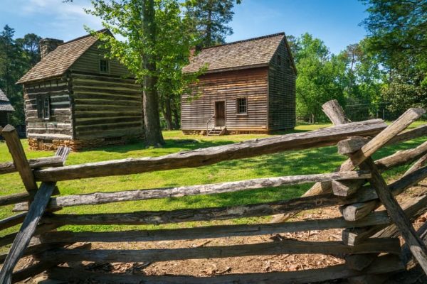 Rustic wooden cabins and a split-rail fence nestled among trees and open grass at Guilford Courthouse National Military Park in Guilford County, NC, offering a glimpse into 18th-century colonial life.
