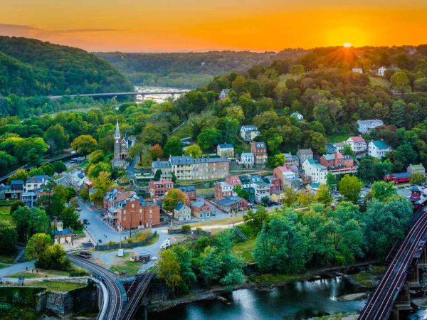 A colorful sunset illuminates the historic town of Harpers Ferry in Jefferson County, West Virginia, nestled among rolling green hills and overlooking a winding river.
