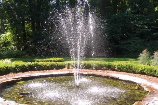 A circular stone fountain spraying water high into the air, surrounded by neatly trimmed bushes and trees in full summer bloom at the Greensboro Arboretum in Guilford County, NC.