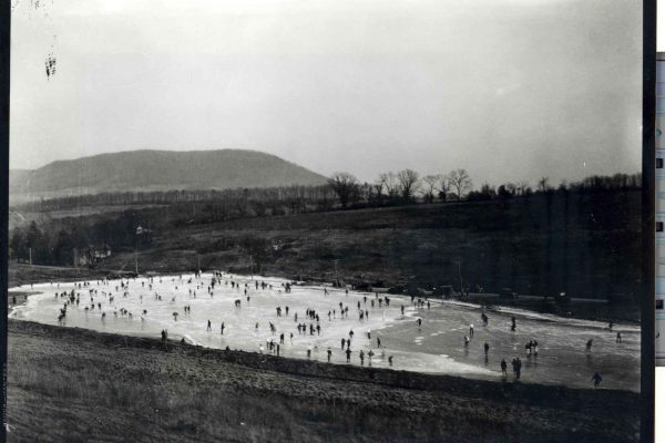 A vintage photo of people ice skating at the Duck Pond near Mount Nittany in Centre County, framed by hills and trees.