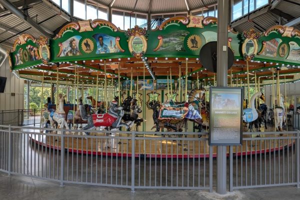 Another angle of the Rotary Club of Greensboro Carousel in Guilford County, NC, showing intricately painted panels, a red NC State wolf carousel figure, and informational signage about Bennett College. The scene captures the carousel’s historical and cultural design elements.