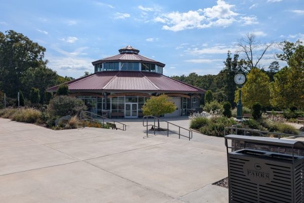 Exterior view of the Rotary Club of Greensboro Carousel building in Guilford County, NC, showing a red-roofed rotunda-style structure surrounded by landscaped greenery, sidewalks, and a classic-style clock pole on a sunny day.