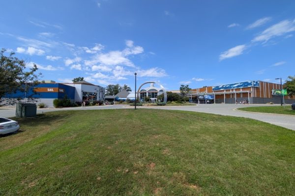Wide-angle view of the Greensboro Science Center in Guilford County, NC, featuring buildings labeled “SAIL” with colorful, modern architecture and a large metal sculpture in the center. Families and children are visible near the entrance on a bright sunny day.